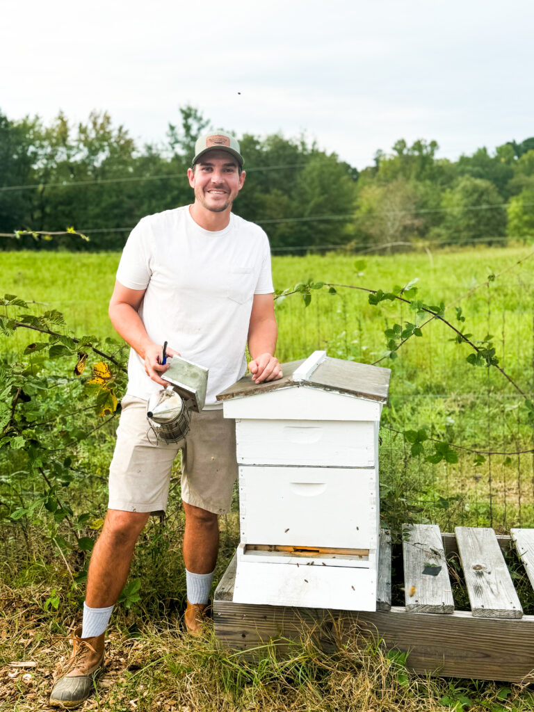Owner and operator James standing next to a enclosed bee hive. There is a field behind him and trees beyond that. Clouds are in the sunny sky.