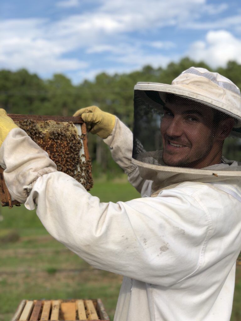The owner and operator James is in his protective gear and holding up a part of a bee hive. There are trees in the background and clouds spread across a blue sky.
